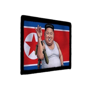 May include: A rectangular patch with a black border featuring a graphic illustration of a person in a white tank top against the North Korean flag. The person is shown sprinkling small green bombs from their hand like salt over their shoulder.