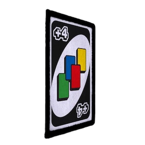 以下が含まれることがあります： 白黒のUnoカードパッチ。白い楕円の中に、緑、赤、青、黄色の4つの長方形が配置されています。カードには白い縁があり、隅には「+4」の文字があります。