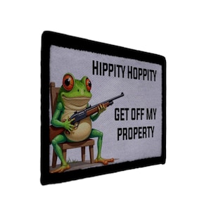 Peut inclure: Écusson avec une grenouille de dessin animé aux yeux rouges tenant un fusil, assise sur une petite chaise en bois. Le patch porte le texte "HIPPITY HOPPITY GET OFF MY PROPERTY" sur fond gris clair avec une bordure noire.