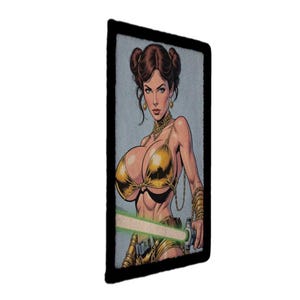 Könnte beinhalten: Ein rechteckiger Aufnäher mit einer Comic-Illustration einer Frau in einem goldenen Bikini, die ein grünes Lichtschwert hält. Das Bild hat einen schwarzen Rand und einen hellblauen Hintergrund.
