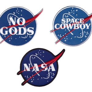 Op de afbeelding: Drie geborduurde patches met een blauwe achtergrond en witte tekst. De patches tonen het NASA-logo met de woorden "NO GODS", "SPACE COWBOY" en "NASA". Elke patch heeft een rood swoosh-ontwerp.