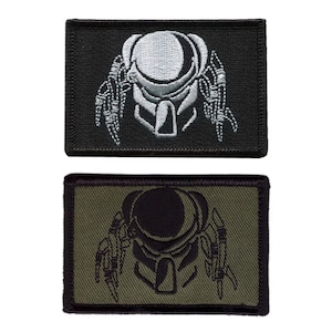 May include: Two rectangular embroidered patches featuring the Predator's face. One patch has a black background with a white design, and the other has an olive green background with a black design. The patches are likely for clothing or gear.