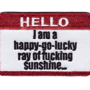 May include: A rectangular patch with a red border and white center. The text "HELLO" is at the top, with the phrase "I am a happy-go-lucky ray of fucking Sunshine..." in black, bold letters.