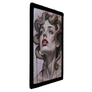May include: A framed portrait of a woman with snakes in her hair, in the style of Medusa. The artwork features a pale complexion, red lips, and blood-like streaks. The snakes are a light brown colour, and the frame is black.