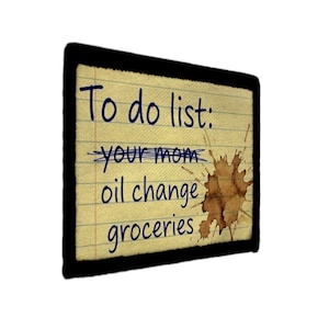 Op de afbeelding: Een rechthoekig notitieblok met een zwarte rand toont een handgeschreven takenlijst. De lijst luidt: "To do list: your mom oil change groceries." De woorden "your mom" zijn doorgestreept en er is een bruine vlek zichtbaar.