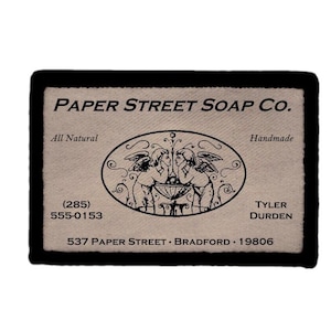 May include: A rectangular fabric label with a black border and beige background. It features the text Paper Street Soap Co., All Natural, Handmade, (285) 555-0153, Tyler Durden, and 537 Paper Street, Bradford, 19806, with a central oval illustration of two cherubs.