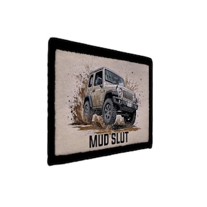 Op de afbeelding: Illustratie van een beige terreinwagen, spetterend door de modder, met de tekst "MUD SLUT" in het zwart. De afbeelding staat tegen een getextureerde, bruine achtergrond, omlijst door een zwarte rand.