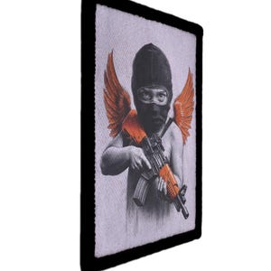 May include: Patch featuring a black and white illustration of a figure wearing a black balaclava and orange wings, holding an orange and black rifle. The patch has a black border and a light gray background.