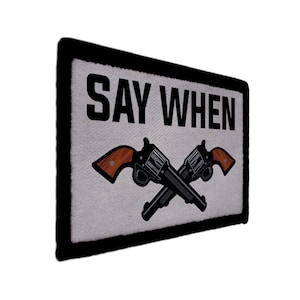 May include: Rectangular patch featuring the text "SAY WHEN" in black. Below the text are two crossed revolvers with brown handles and black barrels. The patch has a white background and a black border.