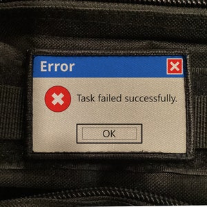 May include: A rectangular patch with a blue top section labelled "Error." Below, a red "X" icon is above the text "Task failed successfully." An "OK" button is at the bottom. The patch is set against a black, textured background.