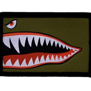 May include: Olive green patch with a shark mouth design, white teeth, red accents, and a black border. The patch has a single eye in the upper left corner. This patch is a military-inspired accessory.
