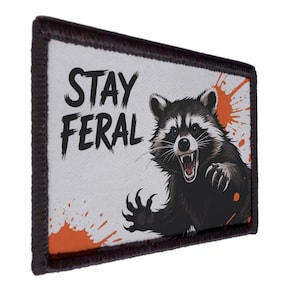 May include: A rectangular patch with a black border and a white background. The patch features a raccoon illustration with the text "STAY FERAL" in black. Orange paint splatters are in the background.