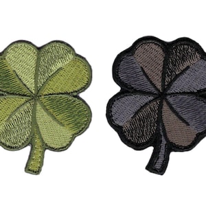 Könnte beinhalten: Zwei gestickte Kleeblatt-Patches. Einer ist grün mit verschiedenen Oliv- und hellgrünen Farbtönen. Der andere ist ein Patchwork aus Grau, Braun und Schwarz. Beide haben einen schwarzen Rand und Stiel.