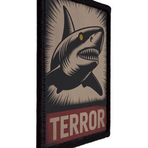 May include: A rectangular patch featuring a menacing shark illustration in black and tan, with a red banner at the bottom that reads "TERROR". The shark has sharp teeth and a yellow eye, set against a background of radiating lines.