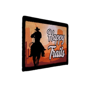 Könnte beinhalten: Ein rechteckiger Aufnäher mit schwarzem Rand zeigt eine Silhouette eines Cowboys auf einem Pferd beim Lasso. Der Hintergrund zeigt eine Wüstenlandschaft in Orange- und Brauntönen. Die Worte "Happy Trails" sind in weißer Schrift geschrieben.