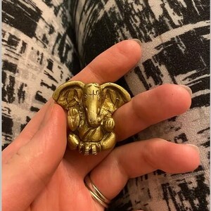 Puede incluir: Un anillo dorado con la forma de la deidad hind&uacute; Ganesha, con detalles intrincados. El anillo presenta al dios con cabeza de elefante en posici&oacute;n sentada. El anillo se sostiene en una mano, con un fondo estampado.