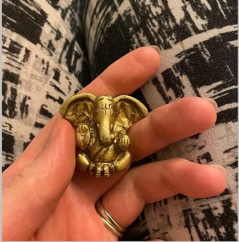 Puede incluir: Un anillo dorado con la forma de la deidad hind&uacute; Ganesha, con detalles intrincados. El anillo presenta al dios con cabeza de elefante en posici&oacute;n sentada. El anillo se sostiene en una mano, con un fondo estampado.
