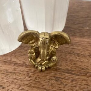 Puede incluir: Una peque&ntilde;a figura de color lat&oacute;n de un dios elefante con grandes orejas y una trompa detallada. La figura est&aacute; sobre una superficie de madera, con dos objetos blancos en el fondo.