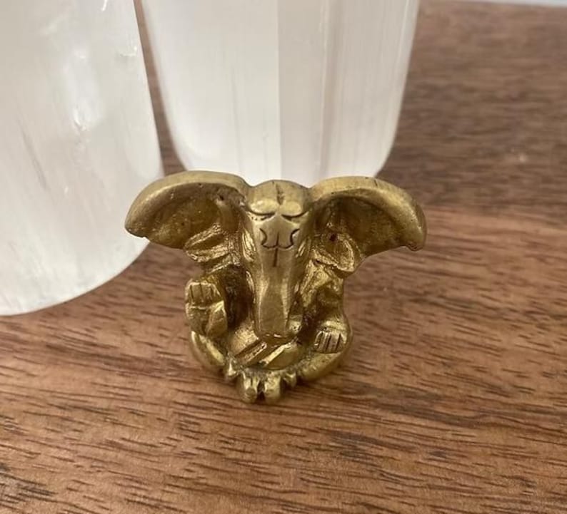 Puede incluir: Una peque&ntilde;a figura de color lat&oacute;n de un dios elefante con grandes orejas y una trompa detallada. La figura est&aacute; sobre una superficie de madera, con dos objetos blancos en el fondo.
