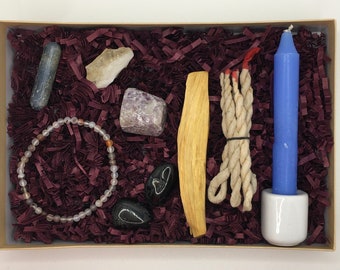 Könnte beinhalten: Eine Geschenkbox gefüllt mit violetten, weißen und schwarzen Kristallen, einem Holzstäbchen Weihrauch, einem weißen Seil und einer blauen Kerze in einem weißen Halter. Die Box ist mit burgunderfarbenem Schredderpapier gefüllt.