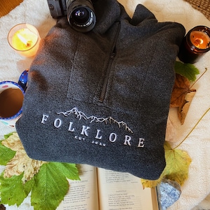 Puede incluir: Una sudadera de forro polar gris oscuro con un logotipo bordado en blanco que dice "FOLKLORE EST. 2020". El logotipo presenta una cordillera de montañas estilizada. La sudadera está sobre una superficie blanca con un libro, una cámara y algunas hojas de otoño.