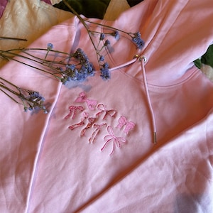 May include: A pink hoodie with a drawstring hood and embroidered pink bows. The hoodie is lying on a white and green surface with dried flowers.