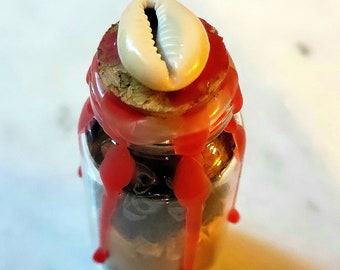 Op de afbeelding: Een kleine glazen pot gevuld met donkere inhoud, afgesloten met een kurk en bekroond met een witte zeeschelp. Rode was druipt langs de zijkanten van de pot, wat een decoratief effect creëert. De pot is waarschijnlijk een decoratief item of een container voor kleine items.