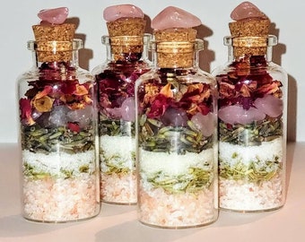 May include: Four clear glass bottles filled with layers of pink salt, white granules, green herbs, and dried flowers. Each bottle is topped with a cork and a pink crystal. The bottles are arranged on a light pink surface.