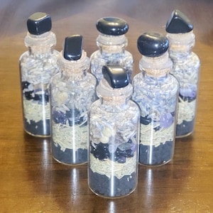 Könnte beinhalten: Sechs kleine Glasflaschen, gefüllt mit Schichten aus dunkelgrauen, grünen und violetten Materialien. Jede Flasche hat einen schwarzen Steinverschluss und einen Korken. Die Flaschen sind auf einer Holzoberfläche angeordnet.