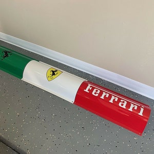 May include: A red, white, and green curved metal sign with the Ferrari logo and the word "Ferrari" in white letters.
