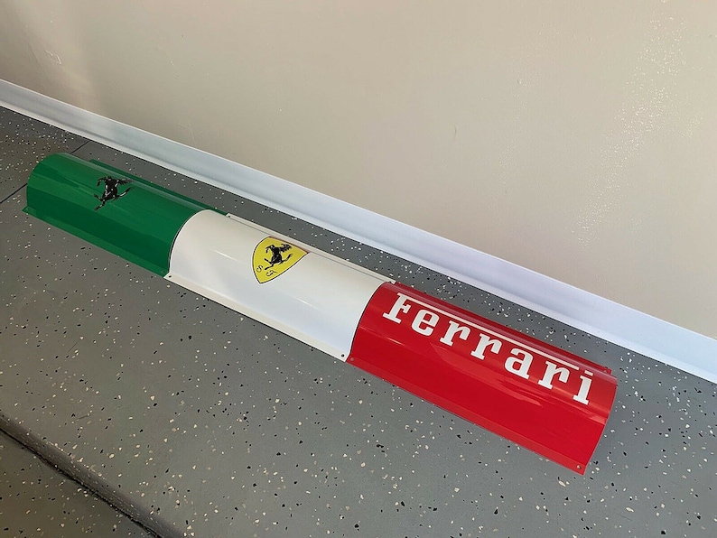 May include: A red, white, and green curved metal sign with the Ferrari logo and the word "Ferrari" in white letters.