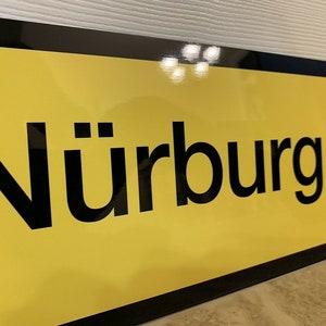 May include: A yellow sign with black lettering that reads "N&uuml;rburgring".