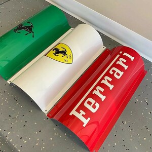 May include: A three-piece metal sign with a green, white, and red design. The green section features a black silhouette of a horse rearing up on its hind legs. The white section features a yellow shield with a black silhouette of a horse rearing up on its hind legs. The red section features the word "Ferrari" in white letters.
