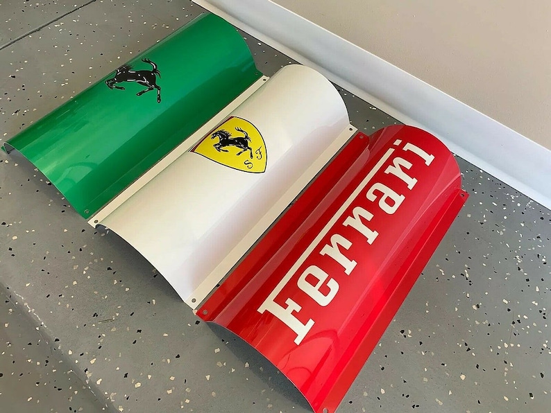 May include: A three-piece metal sign with a green, white, and red design. The green section features a black silhouette of a horse rearing up on its hind legs. The white section features a yellow shield with a black silhouette of a horse rearing up on its hind legs. The red section features the word "Ferrari" in white letters.