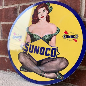 May include: A vintage round metal sign with a yellow background and a blue border. The sign features a pin-up style illustration of a woman wearing a green and white dress and black stockings. The sign also features the text "SUNOCO" in a large, bold font.