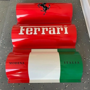 May include: Three red, white, and green metal signs with curved edges. The top sign features a black prancing horse logo and the word "Ferrari". The middle sign says "Ferrari" in white letters. The bottom sign has the words "MODENA" and "ITALIA" in black letters.