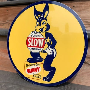 May include: A round yellow and blue metal sign featuring a cartoon bunny wearing a police hat and holding a red and white sign that reads "School Slow Crossing". The bunny is standing on a box that says "Everybody Loves Bunny Enriched Bread".