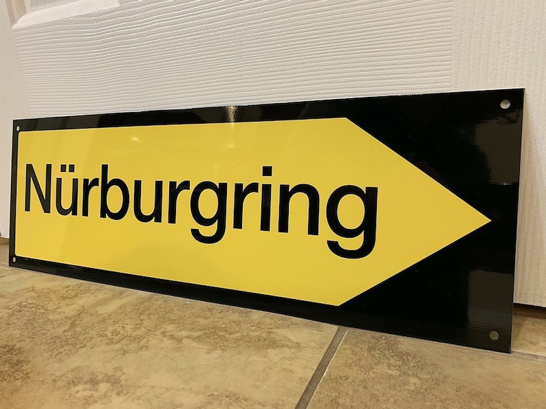 May include: A yellow and black sign with a black arrow pointing to the right. The sign reads "N&uuml;rburgring".
