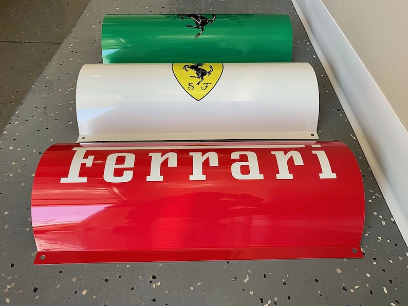 May include: Three red, white, and green cylindrical metal signs with the word "Ferrari" in white letters on the red sign. The white sign has a yellow shield with a black prancing horse logo and the letters "SF" inside the shield. The green sign has a black prancing horse logo.
