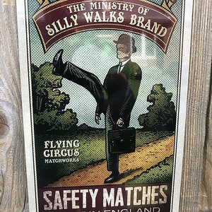Op de afbeelding: Een vintage-stijl illustratie van een man in pak en hoed die loopt met een aktetas. De afbeelding is op een witte achtergrond met een rode banner waarop "The Ministry of Silly Walks Brand" staat. Onder de afbeelding staat een rode banner met de tekst "Safety Matches Made in England".