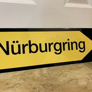 May include: A black and yellow sign with a white arrow pointing to the right. The sign reads "N&uuml;rburgring".