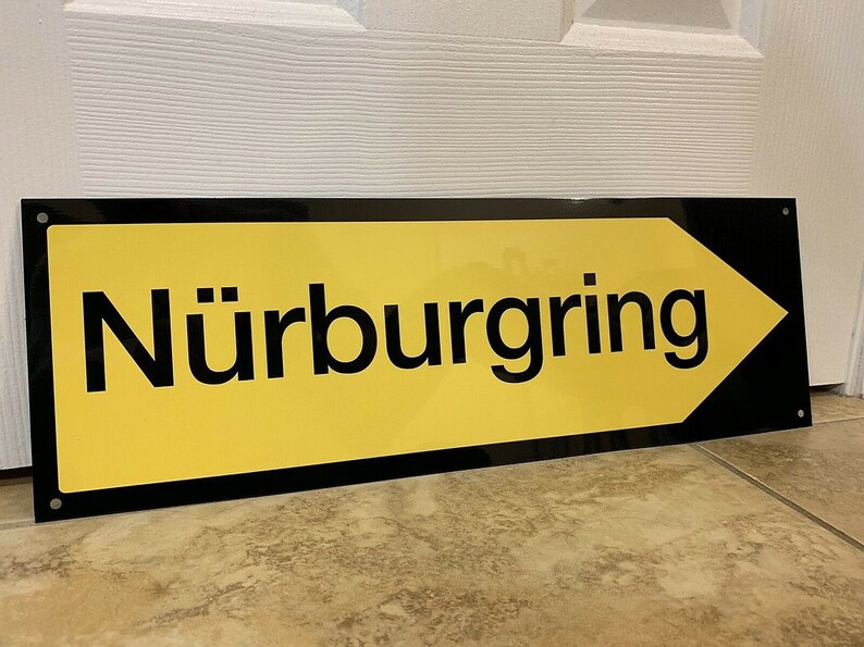 May include: A black and yellow sign with a white arrow pointing to the right. The sign reads "N&uuml;rburgring".