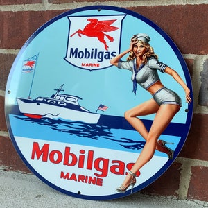 Może przedstawiać: Winietkowy znak Mobiloil Marine z niebieskim tłem i czerwono-białym logo konia. Kobieta w stroju marynarskim stoi na łodzi w wodzie. Na znaku widnieje napis "Mobilgas Marine".