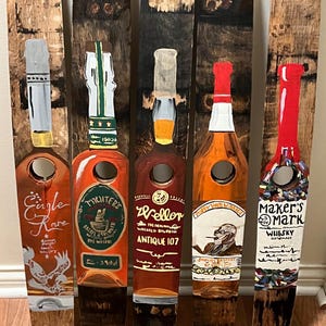 May include: Five wooden bottle stoppers painted to resemble different brands of whiskey. The stoppers are painted with the brand names "Eagle Rare", "Michter's", "Weller", "Poppy John Winkels", and "Maker's Mark".