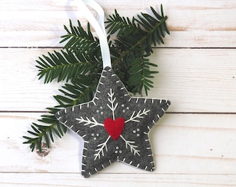 Puede incluir: Un adorno de estrella de fieltro gris con un diseño de copo de nieve blanco y un corazón de fieltro rojo en el centro. El adorno tiene una cinta blanca para colgar.