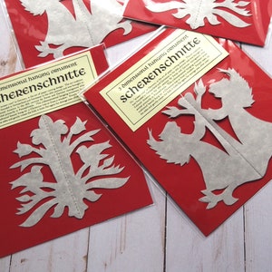 Pode incluir: Duas embalagens vermelhas com recortes de papel branco no interior. As embalagens s&atilde;o etiquetadas "3 dimensional hanging ornament Scherenschnitte".