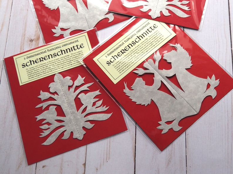 Pode incluir: Duas embalagens vermelhas com recortes de papel branco no interior. As embalagens s&atilde;o etiquetadas "3 dimensional hanging ornament Scherenschnitte".