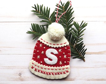 Pode incluir: Um enfeite de Natal de feltro em forma de chapéu de tricô vermelho e branco com um pompom branco no topo. O chapéu tem uma letra "S" branca bordada na frente.