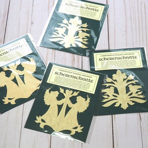 Pode incluir: Quatro pacotes de ornamentos pendurados tridimensionais chamados "Scherenschnitte". Cada pacote cont&eacute;m um design diferente, incluindo anjos, uma &aacute;rvore e uma flor. Os pacotes est&atilde;o sobre uma superf&iacute;cie de madeira clara.