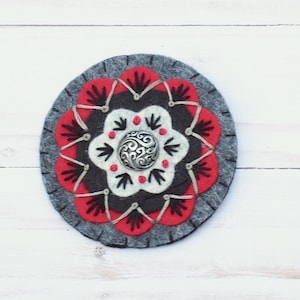 May include: A round felt coaster with a floral design. The coaster features a gray border, red and black petals, and a white center with a silver button. The design is symmetrical and detailed.